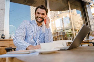 Genç adam dışarıda kafede dizüstü bilgisayarla çalışırken cep telefonuyla konuşuyor.