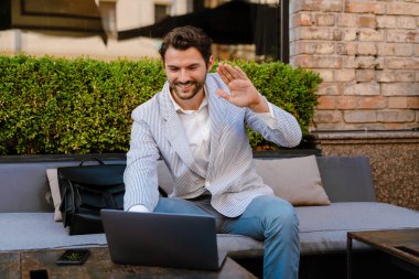 Genç adam dışarıda kafede dizüstü bilgisayarla çalışırken el kol hareketi yapıyor.