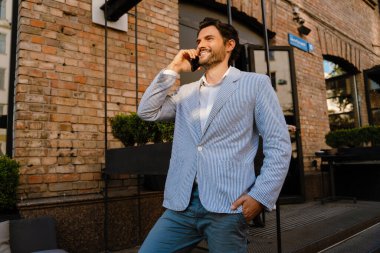 Genç adam dışarıda dururken gülümsüyor ve telefonla konuşuyor.