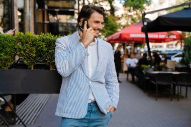 Genç adam dışarıda dururken gülümsüyor ve telefonla konuşuyor.