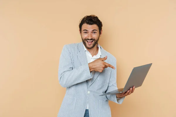 Young white man wearing jacket pointing finger at his laptop isolated over beige background