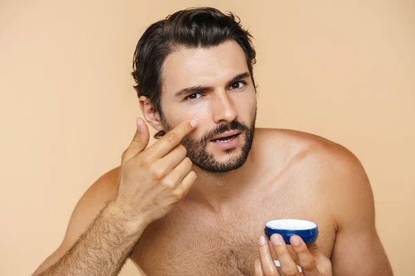 Joven Hombre Semidesnudo Mirando Cámara Mientras Aplica Crema Facial Aislada — Foto de Stock