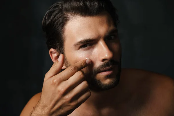 Joven Hombre Semidesnudo Mirando Cámara Mientras Aplica Crema Facial Aislada —  Fotos de Stock