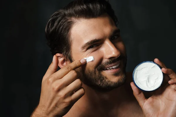 Joven Hombre Semidesnudo Sonriendo Mientras Aplica Crema Facial Aislada Sobre — Foto de Stock