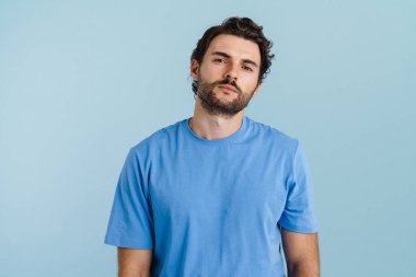 Kıllı saçlı genç adam mavi arka planda izole edilmiş kameraya bakıyor ve poz veriyor