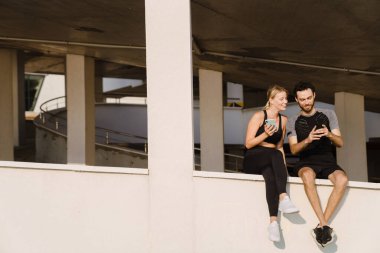 Genç adam ve kadın dışarıda park yerinde gülümsüyor ve cep telefonu kullanıyor.