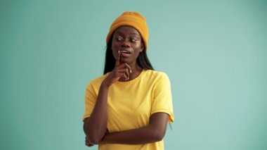 Turkuaz stüdyodaki gülümseyen Afrikalı kadın düşünüyor ve kamerayı işaret ediyor.