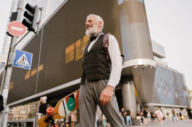 Gri, olgun bir adam, şehir sokaklarında yürürken elinde kaykayla gülümsüyor.