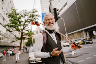 Gri, olgun bir adam, şehir sokağında kaykayla dururken cep telefonu kullanıyor.