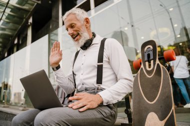 Gri, olgun bir adam şehirde otururken dizüstü bilgisayarla el kol hareketi yapıyor.