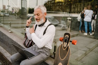 Gri, olgun bir adam şehirde otururken dizüstü bilgisayarla el kol hareketi yapıyor.