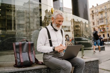 Şehirde otururken dizüstü bilgisayarla çalışırken gülümseyen olgun gri bir adam.