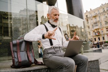 Gri, olgun bir adam şehirde otururken dizüstü bilgisayarla el kol hareketi yapıyor.