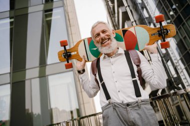 Gri, olgun bir adam, şehir sokağında kaykayla dururken gülümsüyor.