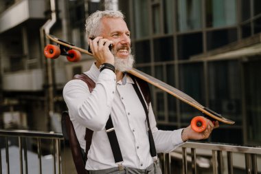 Gri, olgun bir adam, şehir sokağında kaykayla dikilirken cep telefonuyla konuşuyor.