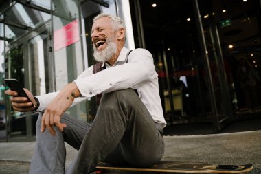 Şehir caddesinde kaykay yaparken cep telefonu kullanan gri, olgun bir adam.