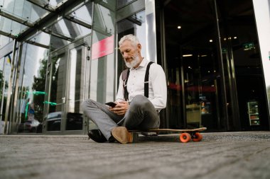 Şehir caddesinde kaykay yaparken cep telefonu kullanan gri, olgun bir adam.