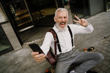 Gri, olgun bir adam, şehirdeki kaykayın üzerinde otururken cep telefonuyla selfie çekiyor.
