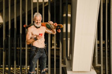 Tişörtsüz, sakallı, olgun bir adam dışarıda kaykay yaparken gülümsüyor.