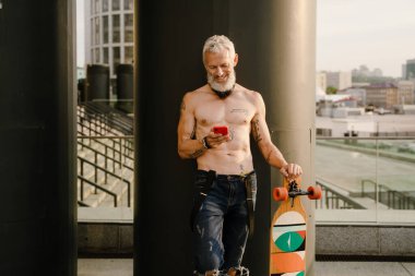 Şehir caddesinde kaykayla gezerken cep telefonu kullanan üstsüz bir adam.