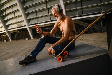 Dışarıda kaykayla otururken cep telefonu kullanan üstsüz, olgun bir adam.