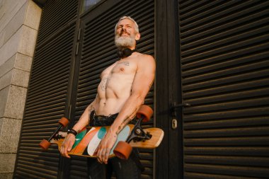 Tişörtsüz, olgun bir adam dışarıda kaykayla dururken gülümsüyor.