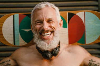 Tişörtsüz, olgun bir adam dışarıda kaykay yaparken gülüyor.