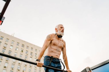 Yetişkin beyaz tişörtsüz adam dışarıda yatay bar kullanarak egzersiz yapıyor.