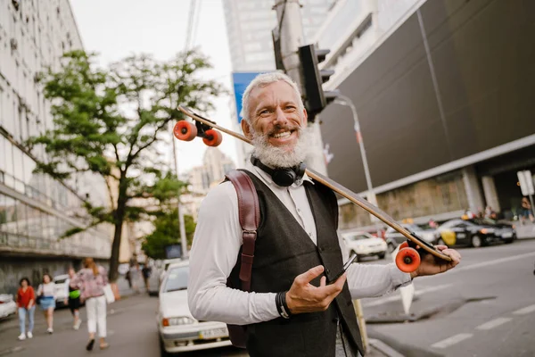 Gri, olgun bir adam, şehir sokağında kaykayla dururken cep telefonu kullanıyor.