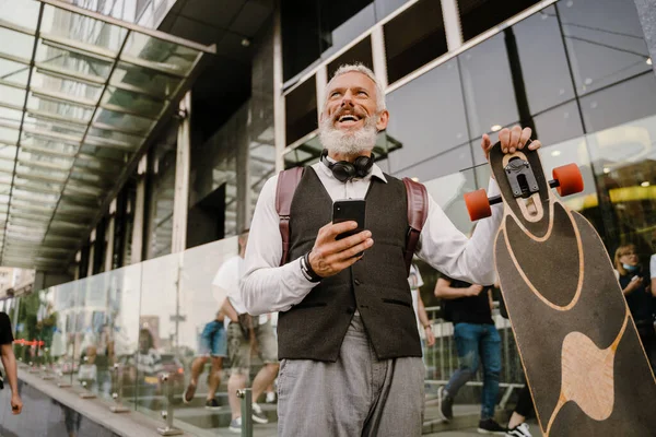 在城市街道上与滑板散步时 灰色的成熟男子用手机 — 图库照片