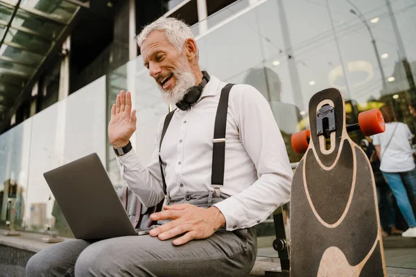 Gri, olgun bir adam şehirde otururken dizüstü bilgisayarla el kol hareketi yapıyor.