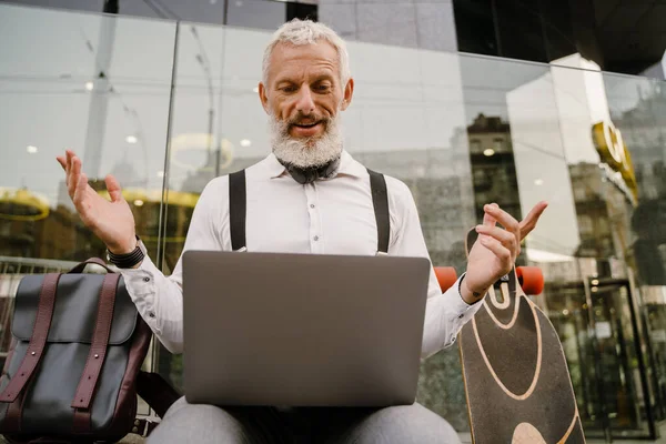 Gri, olgun bir adam şehirde otururken dizüstü bilgisayarla el kol hareketi yapıyor.