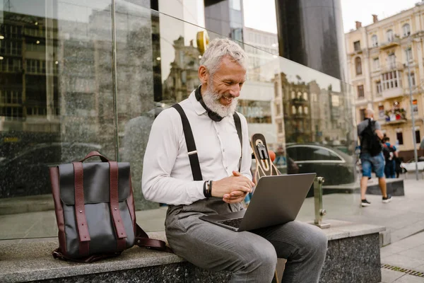 Şehirde otururken dizüstü bilgisayarla çalışırken gülümseyen olgun gri bir adam.