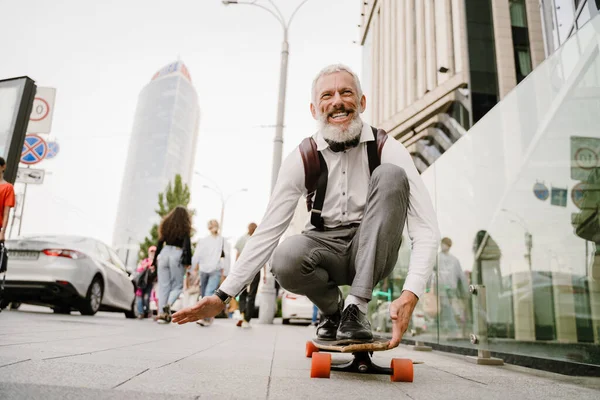 Gri, sakallı, olgun bir adam şehirde kaykay yaparken gülüyor.