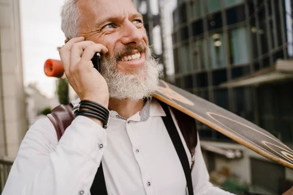 灰色成熟男子站在城市街道上与滑板边用手机交谈 — 图库照片