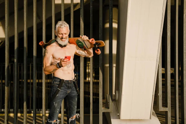 Tişörtsüz, sakallı, olgun bir adam dışarıda kaykay yaparken gülümsüyor.