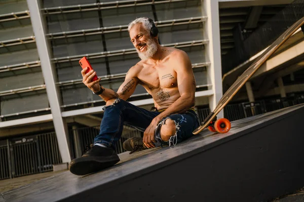 Shirtless Mature Man Using Mobile Phone While Sitting Skateboard Outdoors — стоковое фото