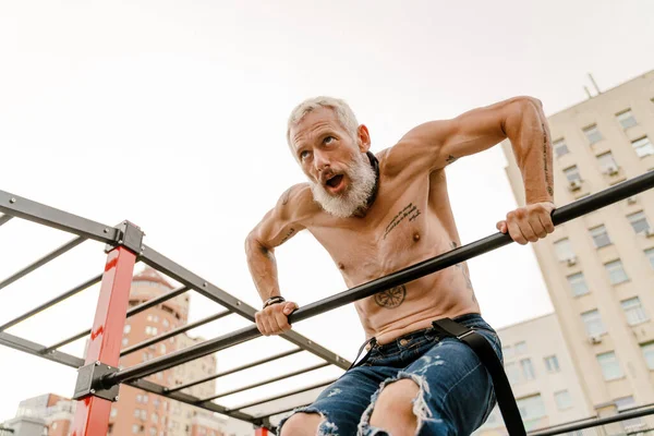 Yetişkin beyaz tişörtsüz adam dışarıda yatay bar kullanarak egzersiz yapıyor.