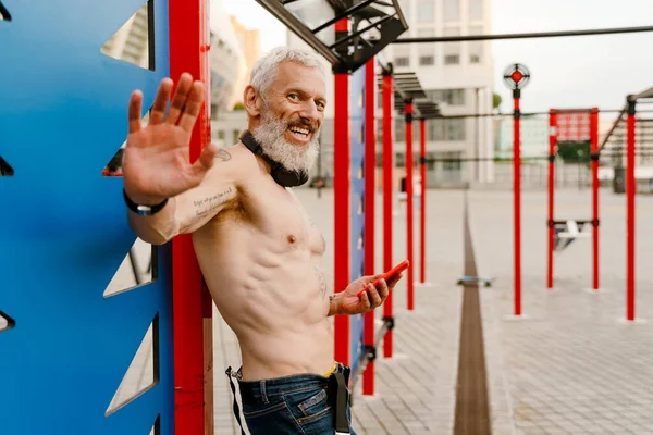 Spor sahasında cep telefonuyla el sallayan ve gülümseyen yetişkin beyaz bir adam.