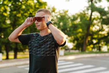Genç beyaz koşucu dışarıda çalışırken güneş gözlüğünü tutuyor.