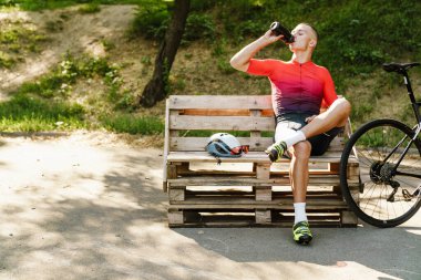 Genç beyaz bisikletçi dışarıda egzersiz yaparken bankta dinlenirken su içiyor.