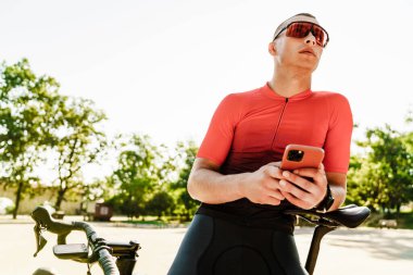 Genç sporcu dışarıda bisikletiyle beklerken cep telefonu kullanıyor.
