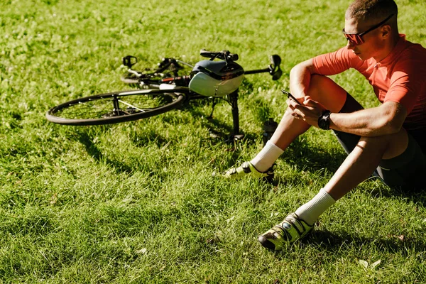 Genç beyaz bisikletçi dışarıda çalışırken çimlerin üzerinde dinlenirken cep telefonu kullanıyor.