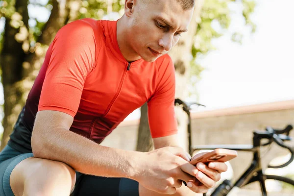 Genç beyaz bisikletçi dışarıda çalışırken bankta dinlenirken cep telefonu kullanıyor.