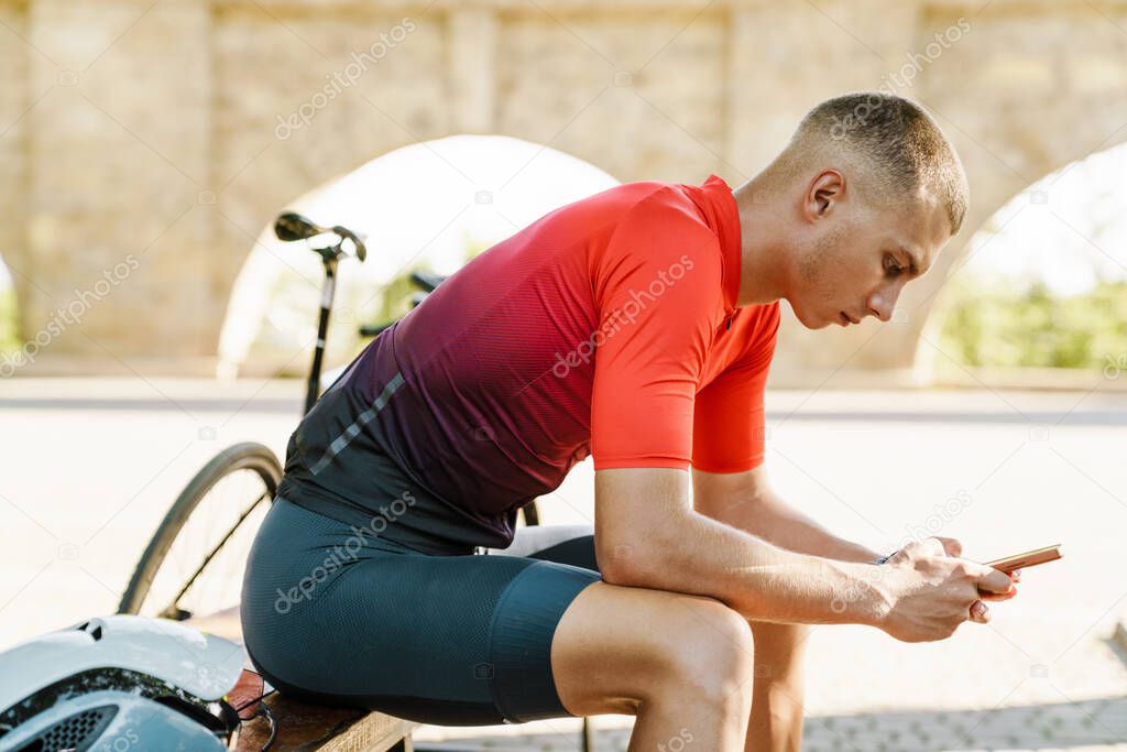 Young white cyclist using cellphone while resting on bench during workout outdoors