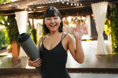 Dışarıda yoga minderiyle dikilirken gülümseyen ve el kol hareketi yapan Asyalı kadın.