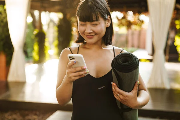 Asian Woman Smiling Using Cellphone While Standing Yoga Mat Outdoors — стоковое фото