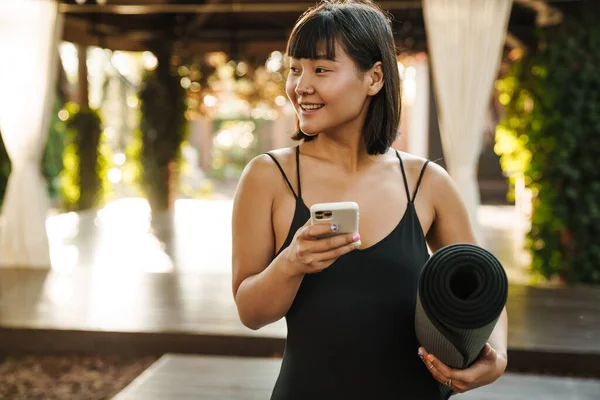 亚洲妇女站在户外带着瑜伽垫 一边笑一边用手机 — 图库照片