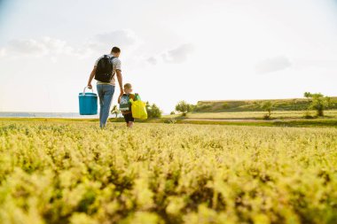 Genç beyaz baba ve oğul yaz tarlasında çantalarla yürüyorlar.