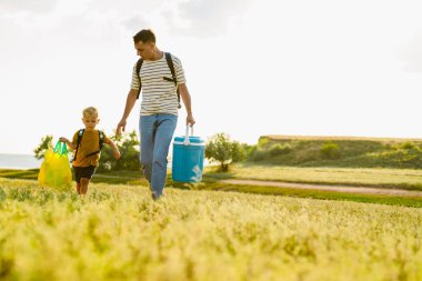 Genç beyaz baba ve oğul yaz tarlasında çantalarla yürüyorlar.